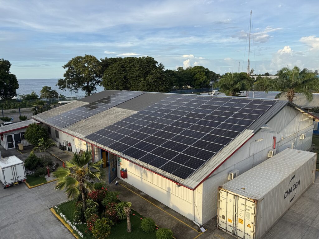 Warehouse with rooftop solar panels near the ocean, surrounded by palm trees, greenery, shipping containers, and a delivery truck.