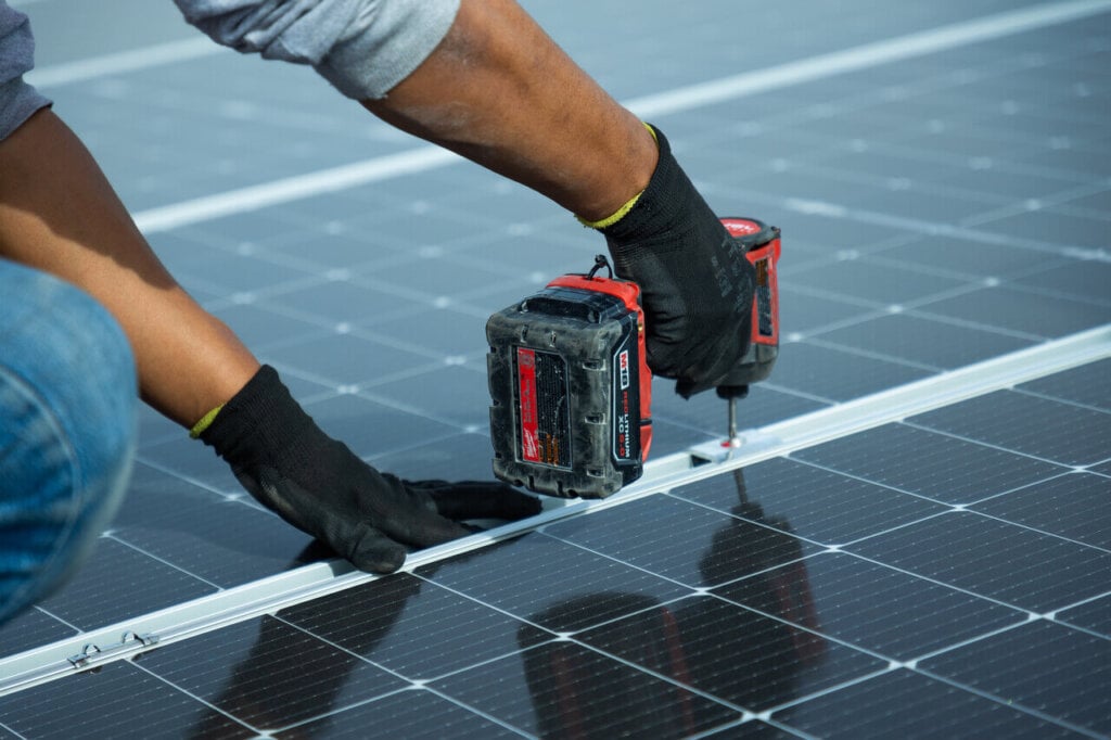Close-up of a solar installer using a power drill to secure the S-5! PVKIT mounting system onto solar panels on a carport roof, demonstrating hands-on installation with gloved hands and precision alignment on photovoltaic modules.