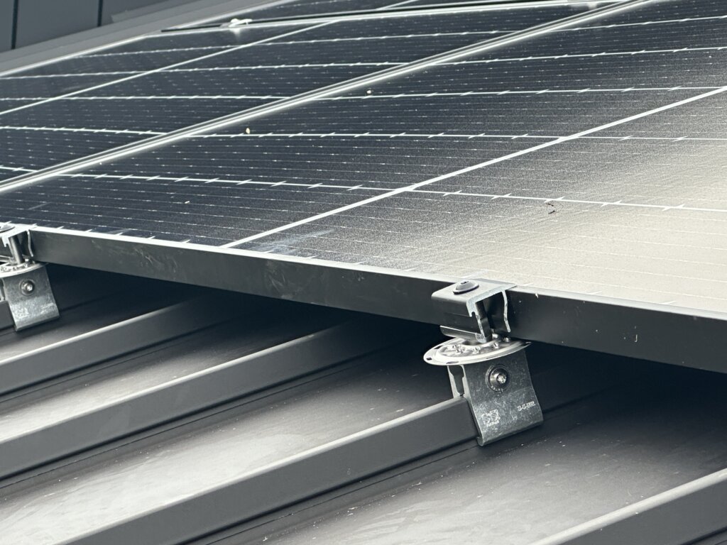 Close-up of solar panel mounting system on a black standing seam metal roof, showing clamps and brackets securing the panel.