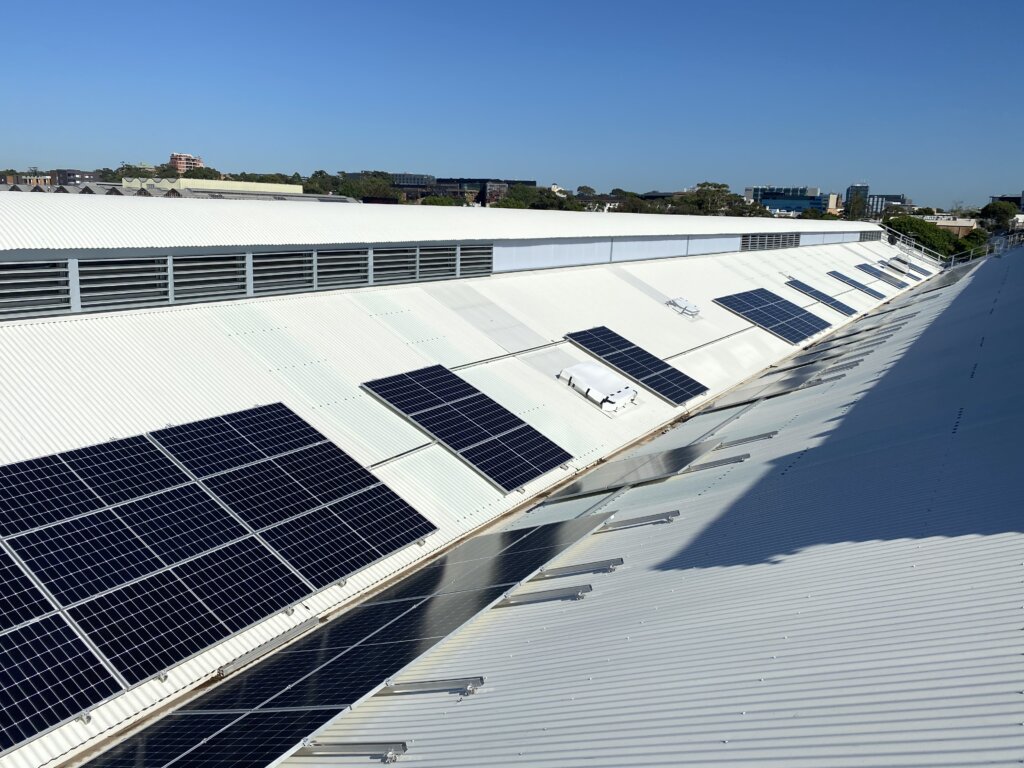Dark solar panel arrays mounted on a large white corrugated metal roof under a clear blue sky.